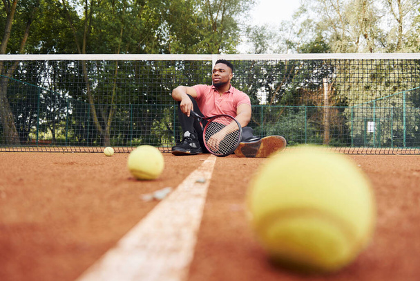 Sits near net and taking a break. African american man in pink shirt sits with tennis racket on the court outdoors. - Foto, imagen