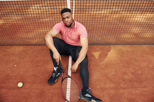 Sits near net and taking a break. African american man in pink shirt sits with tennis racket on the court outdoors. - Foto, afbeelding