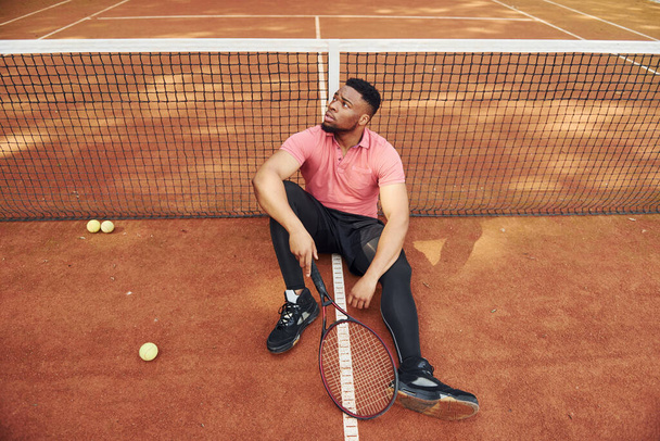 Sits near net and taking a break. African american man in pink shirt sits with tennis racket on the court outdoors. - Photo, Image