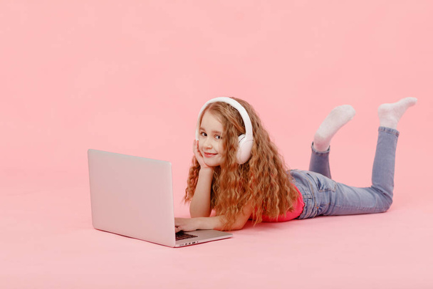Suloinen lapsi tyttö istuu langan vieressä kannettavan tietokoneen. lapsi valkoisissa kuulokkeissa venyttely ja oppii läksyjä tai katsella sarjakuvia, kuunnella musiikkia. - Valokuva, kuva
