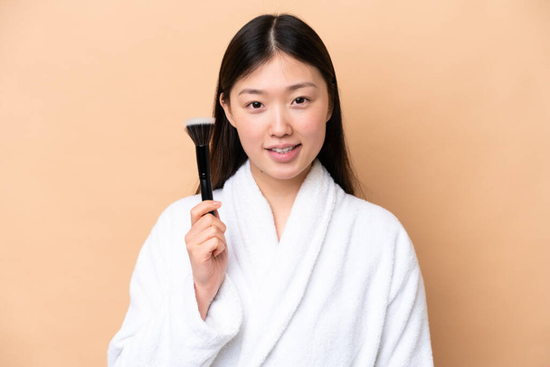 Joven mujer china aislada sobre fondo beige sosteniendo cepillo de maquillaje y whit expresión feliz - Foto, imagen