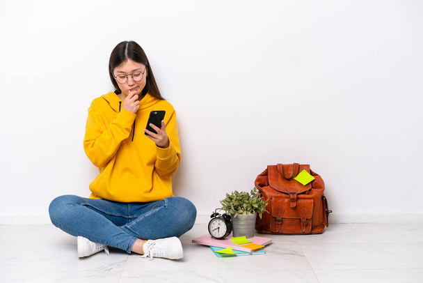 Młoda chińska studentka siedzi na podłodze odizolowana na białej ścianie myśląc i wysyłając wiadomość - Zdjęcie, obraz