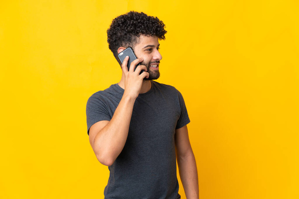 Νεαρός Μαροκινός απομονωμένος σε κίτρινο φόντο κρατώντας μια συνομιλία με το κινητό τηλέφωνο με κάποιον - Φωτογραφία, εικόνα