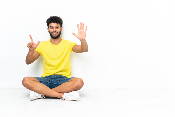 Молодий марокканський вродливий чоловік сидить на підлозі над ізольованим фоном, рахуючи сім пальцями. - Фото, зображення