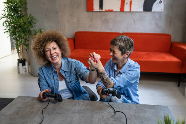 İki Kafkas kadın arkadaş veya kız kardeş ellerinde kumanda paneliyle video oyunu oynuyorlar. Eğleniyorlar. Mutlu olgun kadınlar. - Fotoğraf, Görsel