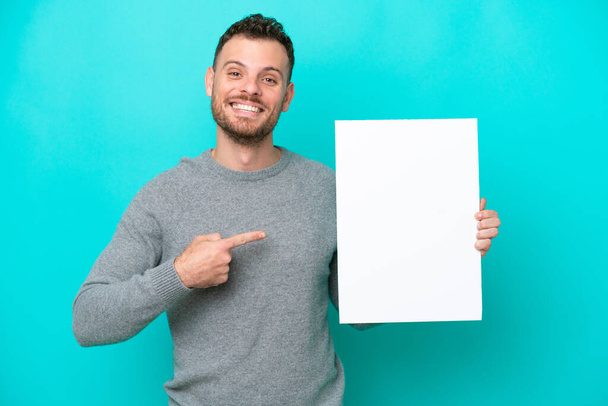 Fiatal brazil férfi, kezében egy üres plakáttal, kék háttérrel, kezében egy üres plakáttal, boldog arckifejezéssel és rámutatva. - Fotó, kép