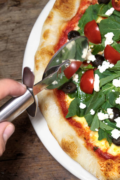 Ready-to-eat pizza with red sauce, mozzarella cheese, arugula, tomato, olive and peasant cheese on a white plate on a wooden background and a pizza cutter - Foto, immagini