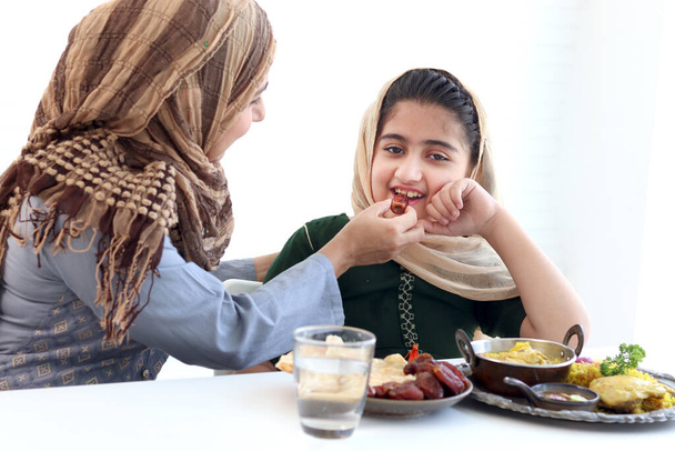 Очаровательная пакистанская мусульманка сидит за кухонным столом, ребенок с хиджабом наслаждается традиционной исламской халяльной пищей с рукой матери на белом фоне, мама кормит ребенка дочь, теплая любовь в семье. - Фото, изображение