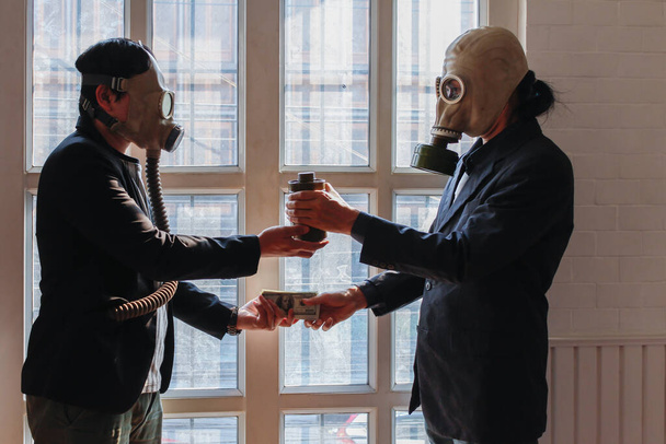 ガスマスクを身に着けている2人の若い男性は、お金のために生物兵器を交換しているテロリストと秘密の組織のグループを表します。有毒ガス漏れの危険を防ぐためにガスマスクを着用するという概念. - 写真・画像