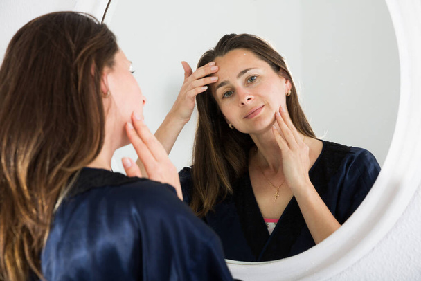Portrait of adult woman looking at mirror and touching face in bedroom - Foto, Imagen