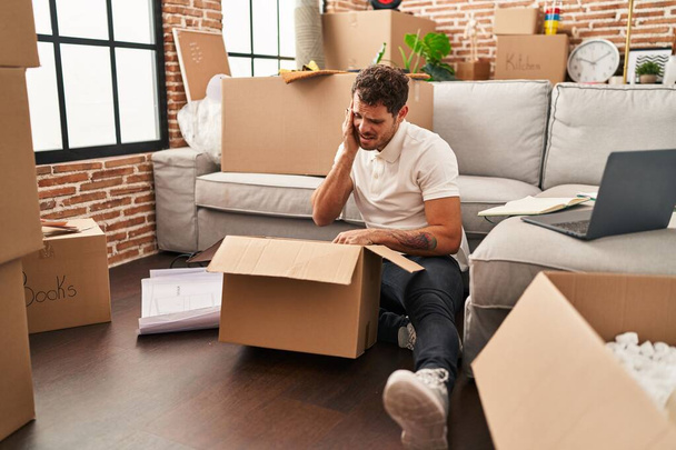 Young hispanic man unboxing package with worry expression at new home - 写真・画像