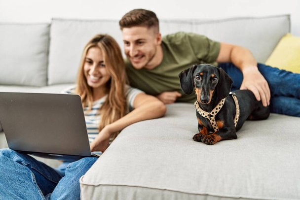 Νεαρό ζευγάρι ισπανόφωνων χαμογελά χαρούμενος χρησιμοποιώντας φορητό υπολογιστή κάθεται στον καναπέ με το σκυλί στο σπίτι. - Φωτογραφία, εικόνα
