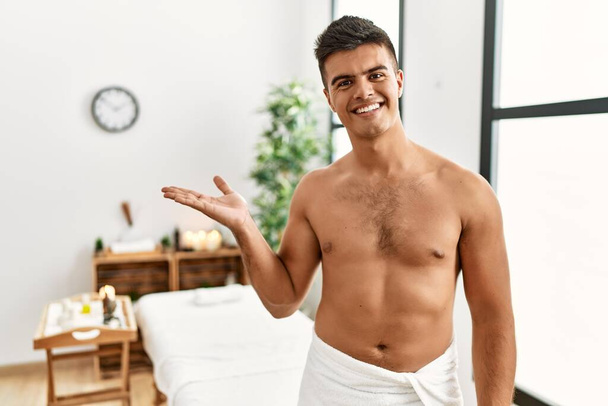Joven hombre hispano de pie sin camisa en el centro de spa sonriendo alegre presentación y señalando con la palma de la mano mirando a la cámara.  - Foto, imagen