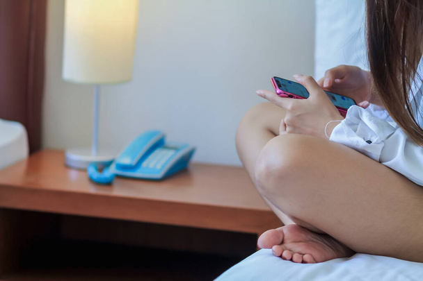 Μικρή Ασιάτισσα γυναίκα χαλαρώνει στο κρεβάτι το πρωί και χρησιμοποιεί το έξυπνο τηλέφωνό της συνδεδεμένο στο Διαδίκτυο για να ελέγξει τα μηνύματα κοινωνικής δικτύωσης μέσω μιας εφαρμογής smartphone. - Φωτογραφία, εικόνα