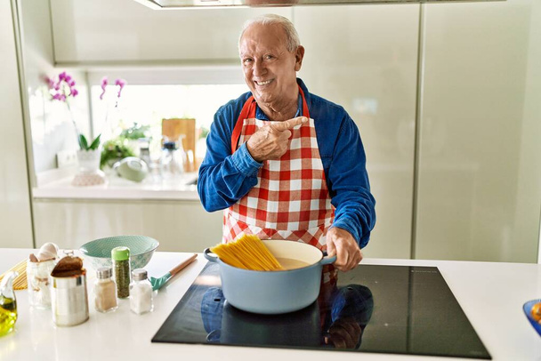 Senior man met grijs haar koken spaghetti thuis keuken lachend gelukkig wijzend met hand en vinger naar de zijkant  - Foto, afbeelding