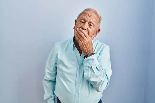Hombre mayor con el pelo gris de pie sobre fondo azul aburrido bostezo cansado cubriendo la boca con la mano. inquietud y somnolencia.  - Foto, imagen