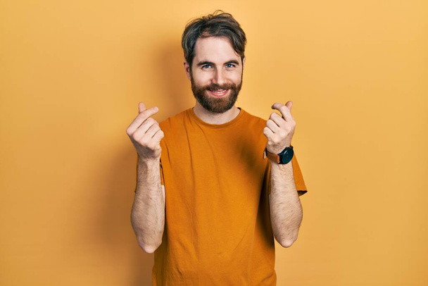 Homme caucasien avec barbe vêtu d'un t-shirt jaune décontracté faisant geste d'argent avec les mains, demandant un paiement de salaire, entreprise millionnaire  - Photo, image