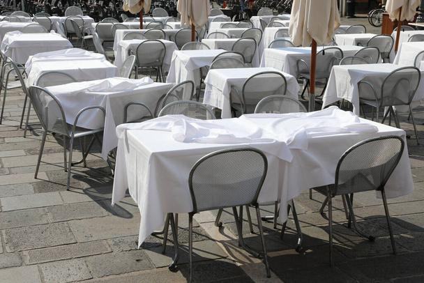 μικρά τραπέζια με λευκό τραπεζομάντηλο και άδειες καρέκλες στην πλατεία της ευρωπαϊκής πόλης χωρίς ανθρώπους λόγω της οικονομικής κρίσης - Φωτογραφία, εικόνα