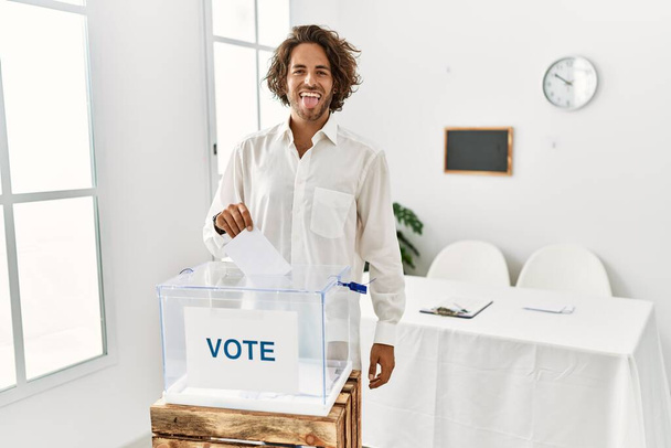 Young hispanic man voting putting envelop in ballot box sticking tongue out happy with funny expression. emotion concept.  - Photo, Image