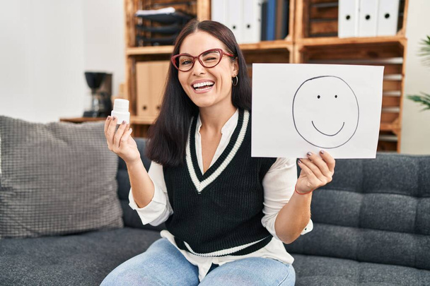 Młoda Latynoska kobieta pracuje nad depresją trzymając szczęśliwe emocje na papierze i pigułki mrugając patrząc w kamerę z seksowną ekspresją, wesołą i szczęśliwą twarzą.  - Zdjęcie, obraz