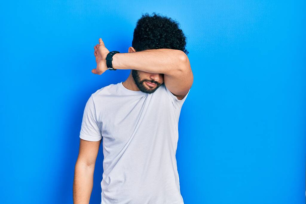 Young arab man with beard wearing casual white t shirt covering eyes with arm, looking serious and sad. sightless, hiding and rejection concept  - Fotografie, Obrázek