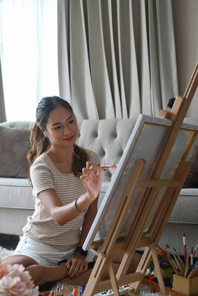 Pretty young woman painting a picture on canvas in bright living room. - Photo, image