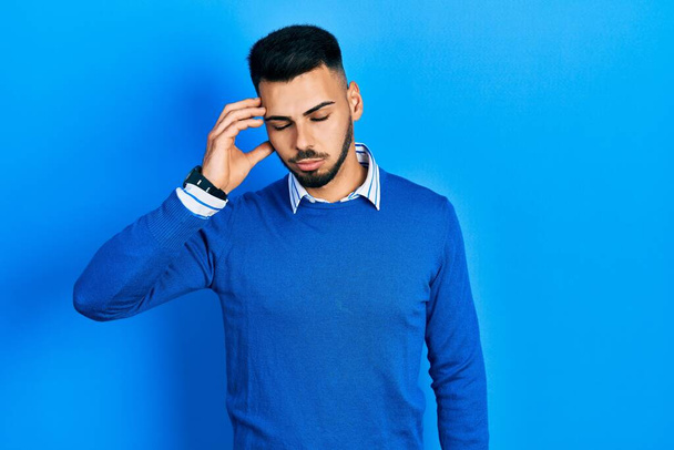 Jonge Spaanse man met baard draagt casual blauwe trui bezorgd en gestrest over een probleem met de hand op het voorhoofd, nerveus en angstig voor crisis  - Foto, afbeelding