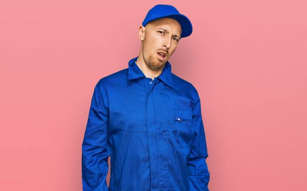Bald man with beard wearing builder jumpsuit uniform in shock face, looking skeptical and sarcastic, surprised with open mouth  - Fotoğraf, Görsel