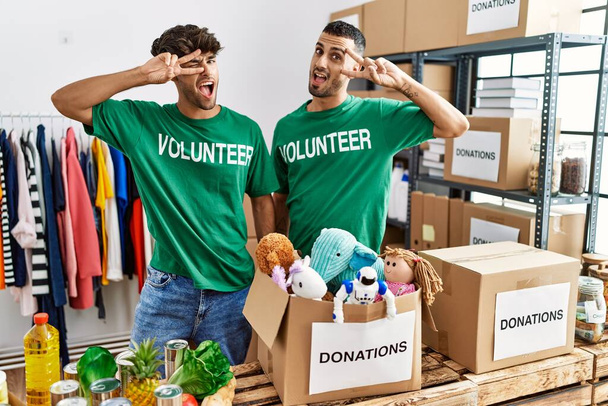 Young gay couple wearing volunteer t shirt at donations stand doing peace symbol with fingers over face, smiling cheerful showing victory  - Photo, Image