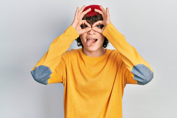 Un joven guapo usando un sombrero de lana haciendo un buen gesto como binoculares sacando la lengua, ojos mirando a través de los dedos. expresión loca.  - Foto, imagen