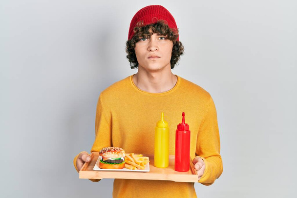 Красивый молодой человек ест вкусный классический бургер с кетчупом и горчицей скептик и нервный, хмурясь расстроен из-за проблемы. отрицательный человек.  - Фото, изображение