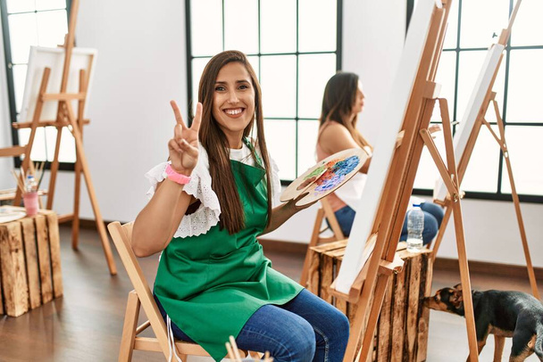 Jonge Latijns-Amerikaanse kunstenares die schildert op doek in een kunststudio, wijzend naar jou en de camera met vingers, positief en vrolijk lachend  - Foto, afbeelding
