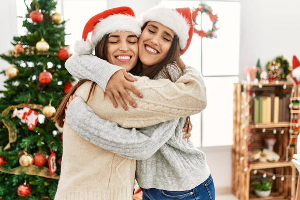 Dos mujeres sonriendo confiadas abrazándose de pie junto al árbol de Navidad en casa - Foto, imagen