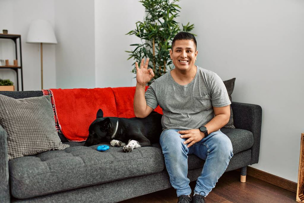 Young latin man and dog sitting on the sofa at home smiling positive doing ok sign with hand and fingers. successful expression.  - Fotó, kép