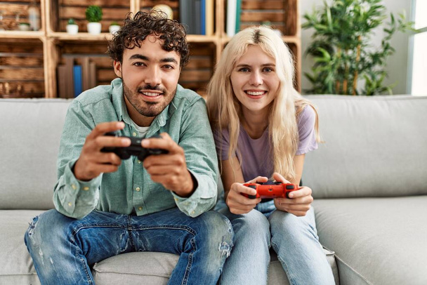 Νεαρό ζευγάρι χαμογελά ευτυχισμένος παίζοντας βιντεοπαιχνίδι στο σπίτι. - Φωτογραφία, εικόνα