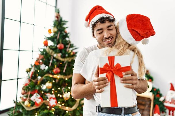 Νεαρό ζευγάρι φοράει χριστουγεννιάτικο καπέλο αγκαλιάζει και κρατώντας δώρο στο σπίτι. - Φωτογραφία, εικόνα