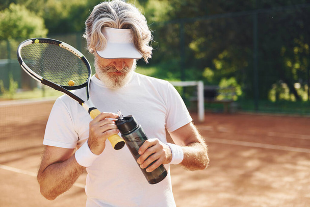 Taking a break and drinking water. Senior modern stylish man with racket outdoors on tennis court at daytime. - Foto, Imagem