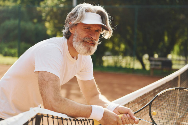 Senior modern stylish man with racket outdoors on tennis court at daytime. - Фото, изображение