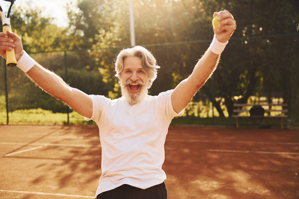 Celebrating victory. Senior modern stylish man with racket outdoors on tennis court at daytime. - Zdjęcie, obraz