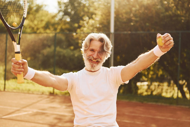 Celebrating victory. Senior modern stylish man with racket outdoors on tennis court at daytime. - Foto, afbeelding