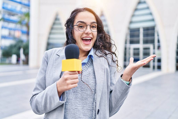Junge brünette Frau hält Reportermikrofon und feiert Leistung mit glücklichem Lächeln und Siegerausdruck mit erhobener Hand  - Foto, Bild