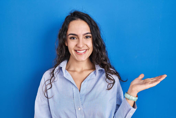 Nuori brunette nainen seisoo sinisellä taustalla hymyilevä iloinen esittelee ja osoittaa kämmenellä katsoen kameraa.  - Valokuva, kuva