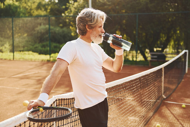 Taking a break and drinking water. Senior modern stylish man with racket outdoors on tennis court at daytime. - Photo, image