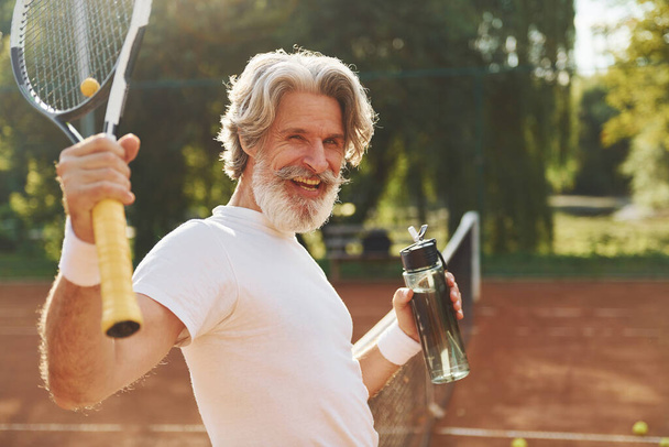 Prendre une pause et boire de l'eau. Homme élégant moderne senior avec raquette à l'extérieur sur un court de tennis le jour. - Photo, image