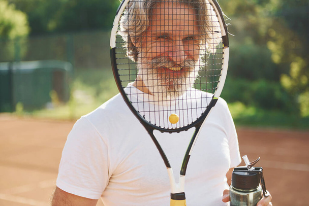 Перерыв и питьевая вода. Старший современный стильный мужчина с ракеткой на открытом воздухе на теннисном корте днем. - Фото, изображение