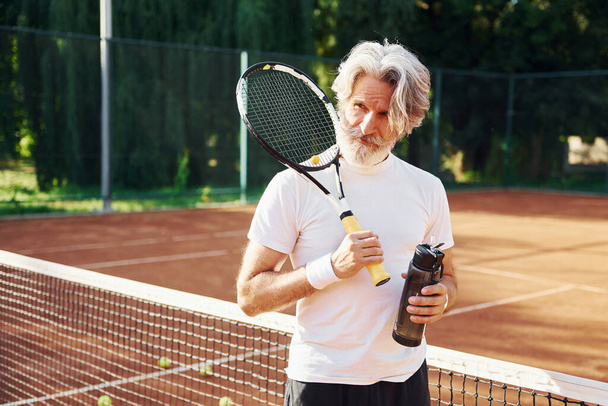 Holding bottle of water. Senior modern stylish man with racket outdoors on tennis court at daytime. - 写真・画像