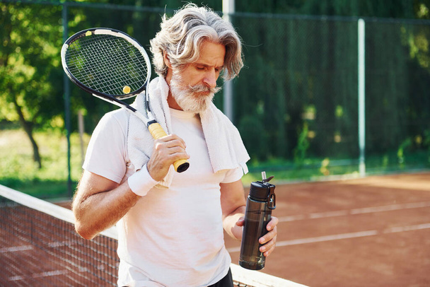 Holding bottle of water. Senior modern stylish man with racket outdoors on tennis court at daytime. - Photo, Image