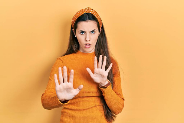Joven adolescente morena vistiendo suéter amarillo casual alejando las palmas de las manos mostrando rechazo y negación con miedo y expresión repugnante. parada y prohibido.  - Foto, imagen