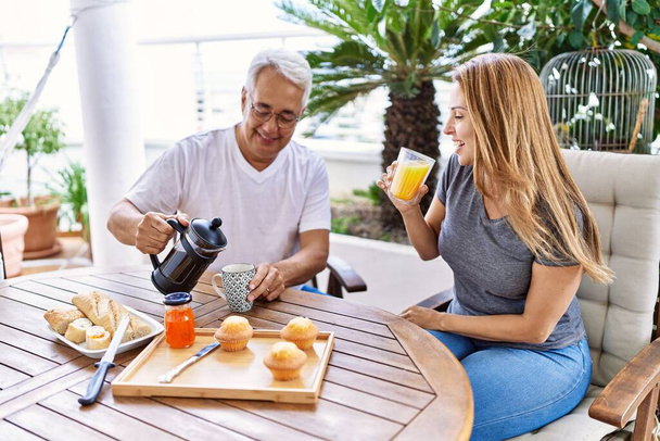 Μεσήλικες ισπανόφωνοι ζευγάρι χαμογελάνε ευτυχισμένοι τρώγοντας πρωινό στη βεράντα. - Φωτογραφία, εικόνα