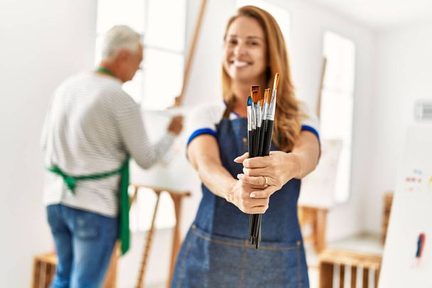 Dos artistas de mediana edad sonriendo feliz pintura en el estudio de arte. Mujer de pie sosteniendo pinceles. - Foto, Imagen